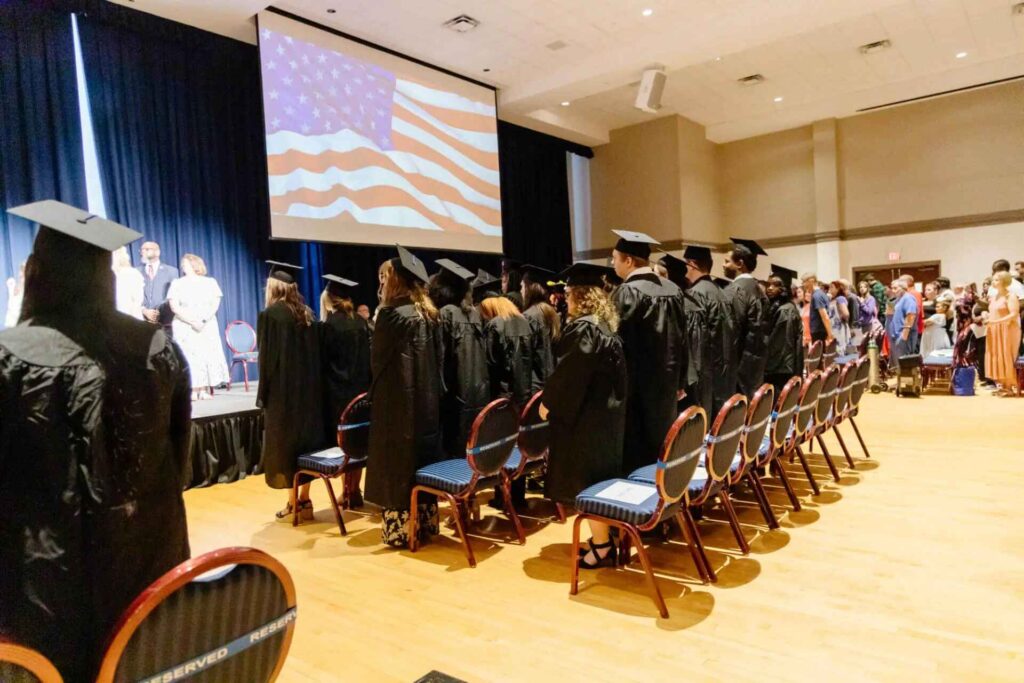 Graduates and attendees stand and face a large screen displaying the American flag during the national anthem. Everyone is dressed formally, with students in caps and gowns and guests filling the audience in the background.