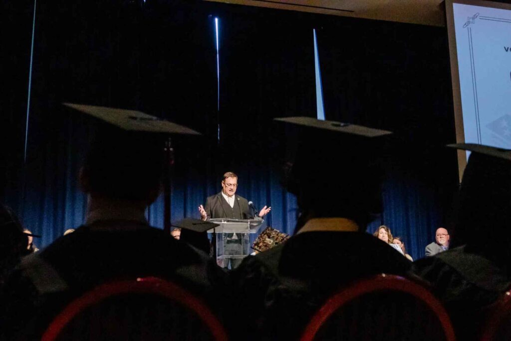A graduation speaker stands behind a clear podium addressing the graduates seated in the foreground. The scene is dimly lit with dark curtains and the graduates’ caps partially visible as silhouettes.